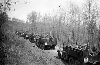 Archival photograph of Hungarian troops riding in an extended convoy of open military trucks on a rural road.