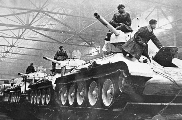 Archival photograph of Soviet T-34 tanks and their crews inside a factory; the archive dates the photograph to 1942.